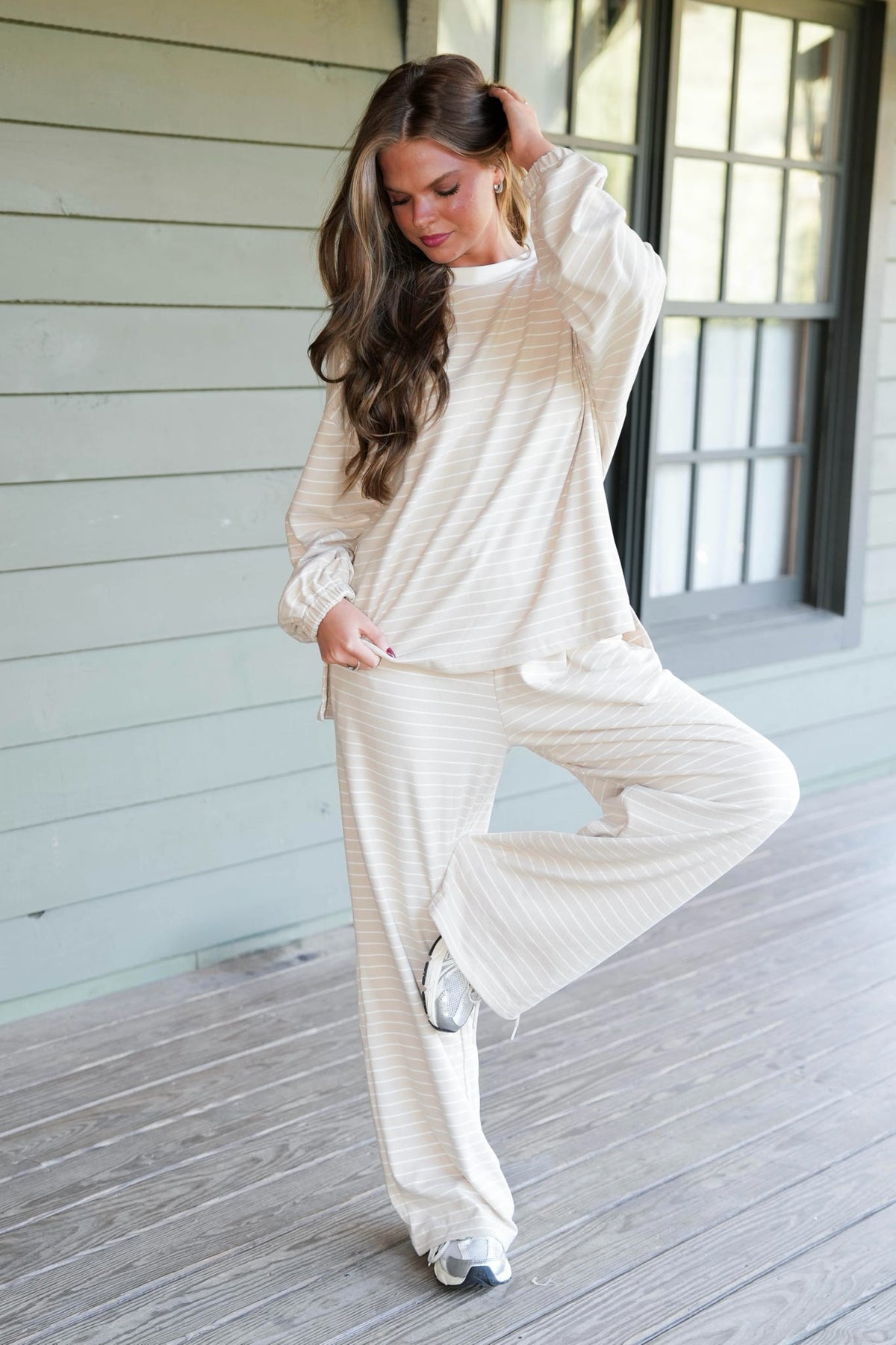 A woman modeling a matching cream and white striped lounge set consisting of a long-sleeve top and wide-leg pants, standing on a wooden deck in front of a house with light green siding.
