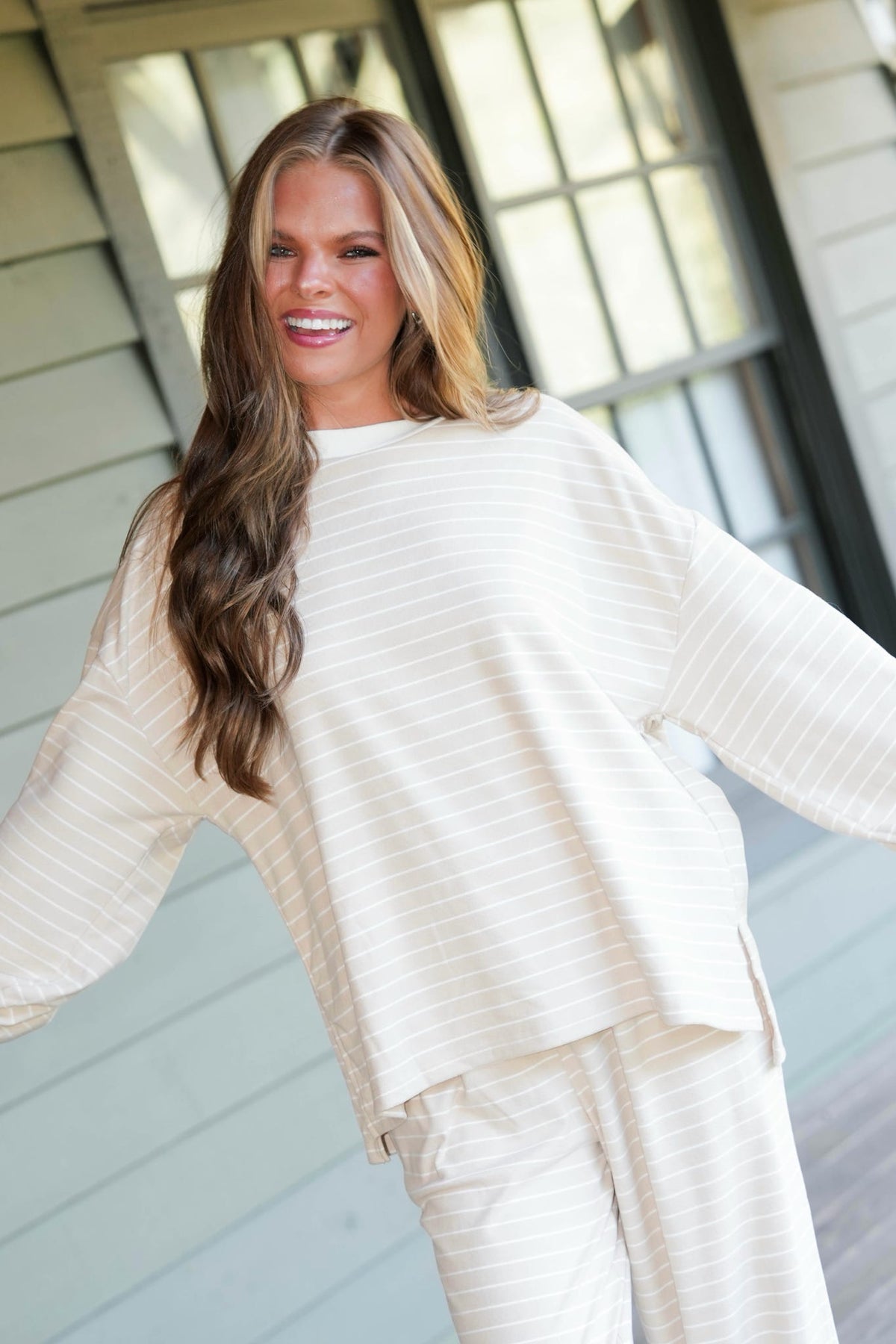 A woman wearing a matching cream and white striped lounge set with a long-sleeve top and pants, standing on a porch with her arms outstretched.