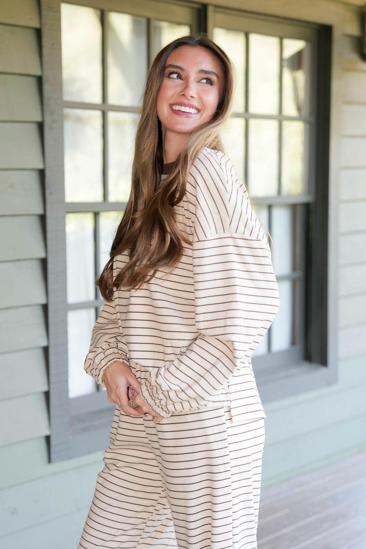 A woman wearing a beige and white striped lounge set stands on a porch in front of a gray-framed window.