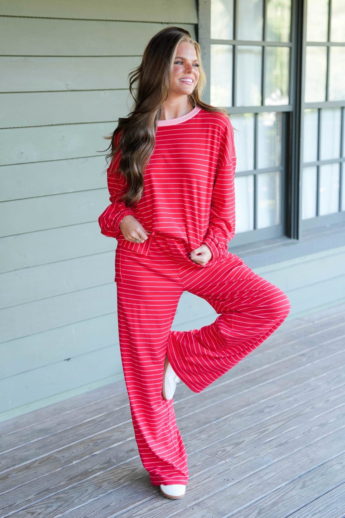A woman modeling a matching red and white striped lounge set consisting of a long-sleeve top and wide-leg pants, standing on a wooden porch.