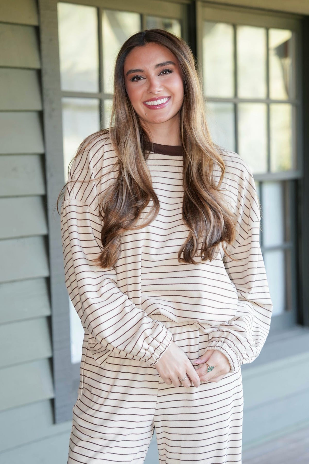 A woman with long brown hair smiling while wearing a beige and brown horizontally striped long-sleeve pajama set, standing on a porch in front of a window.