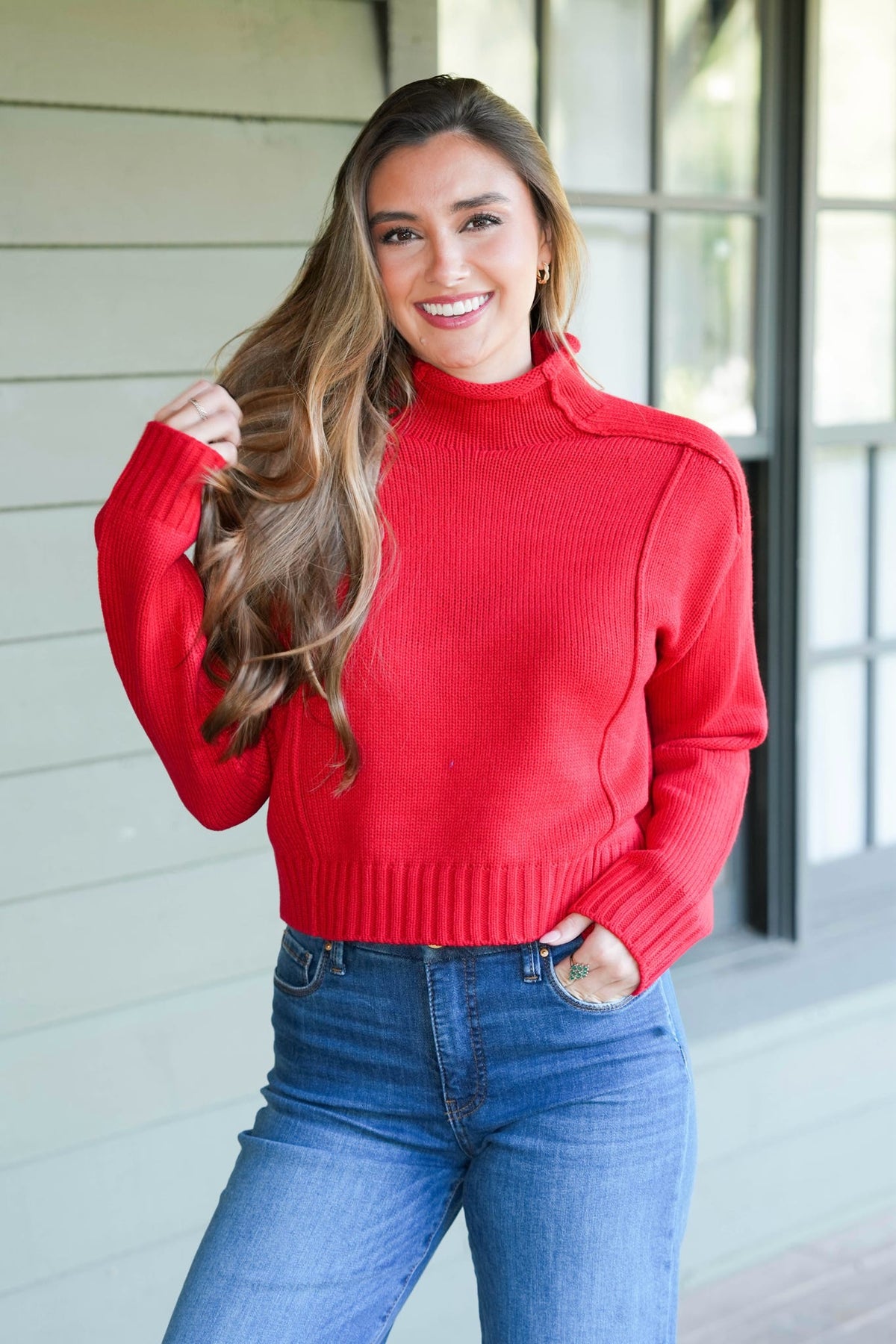 A smiling woman with long brown hair models a bright red ribbed mock neck sweater with shoulder details, paired with blue jeans, standing in front of a light-colored house exterior.