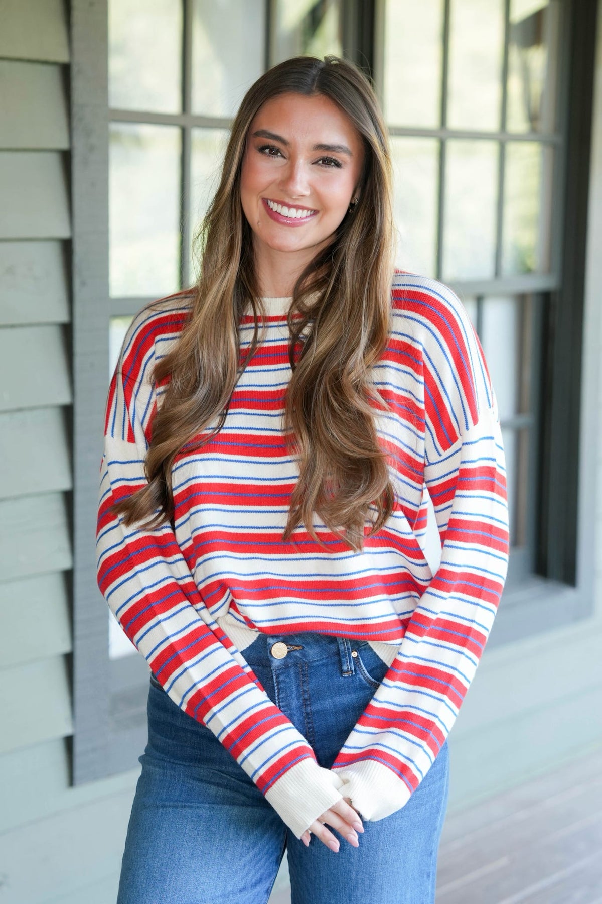 A smiling woman with long brown hair stands on a porch wearing a red, white, and blue striped long-sleeve knit top with blue jeans.