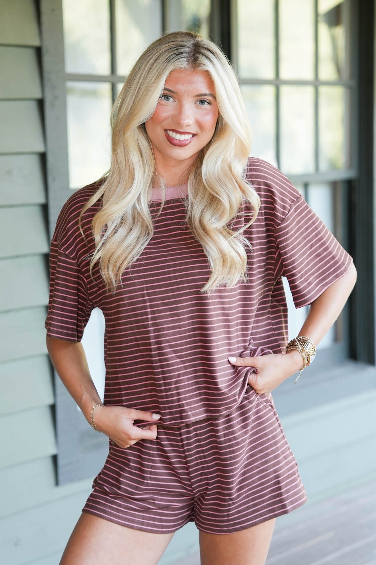 A smiling blonde woman wearing a short-sleeve, crewneck striped romper in a dusty rose and white color combination, standing in front of a house porch with windows.