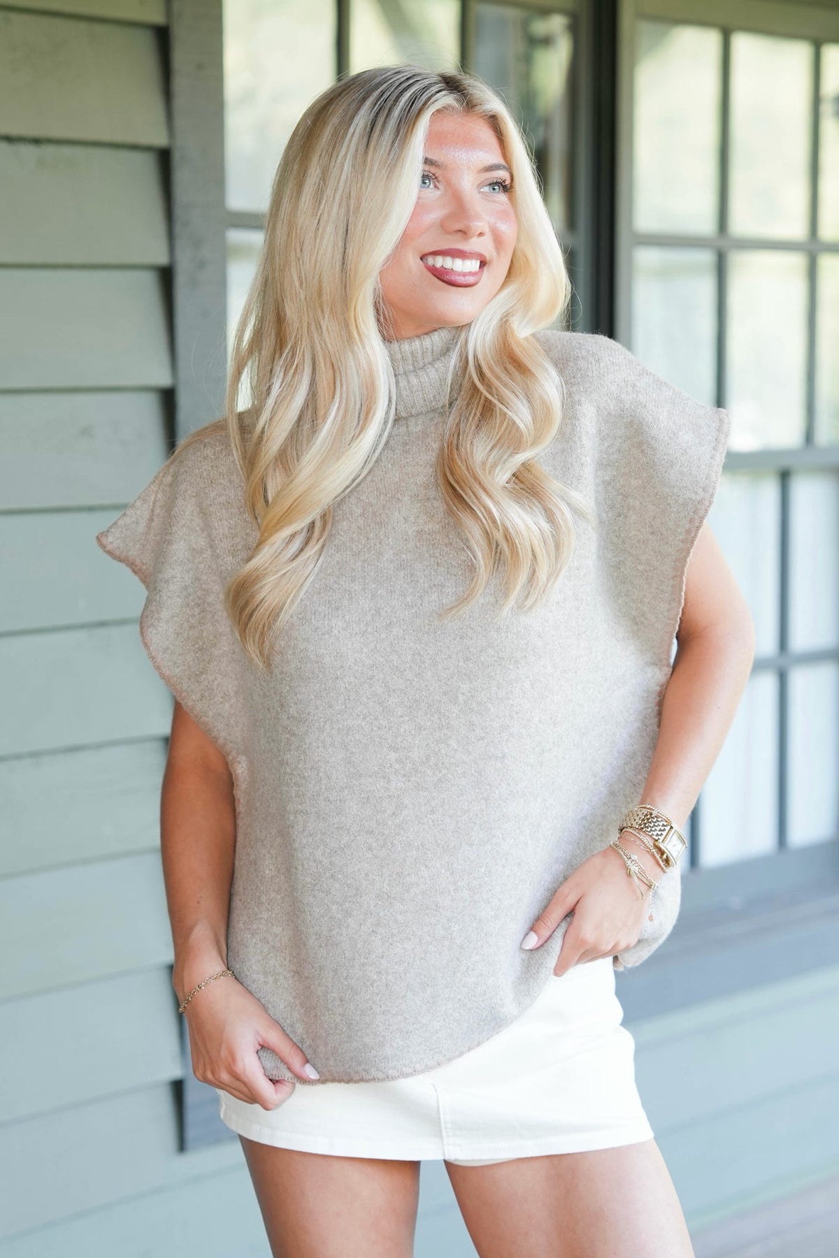 A smiling blonde woman wearing a light beige knit mock-neck top with wide short sleeves and white shorts, standing in front of a gray house exterior.