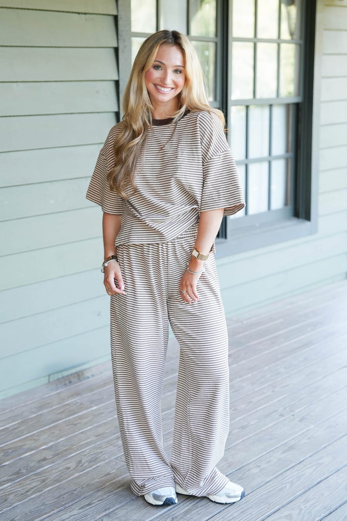 A woman stands on a wooden porch wearing a matching beige and white striped lounge set consisting of a short-sleeve top and wide-leg pants.