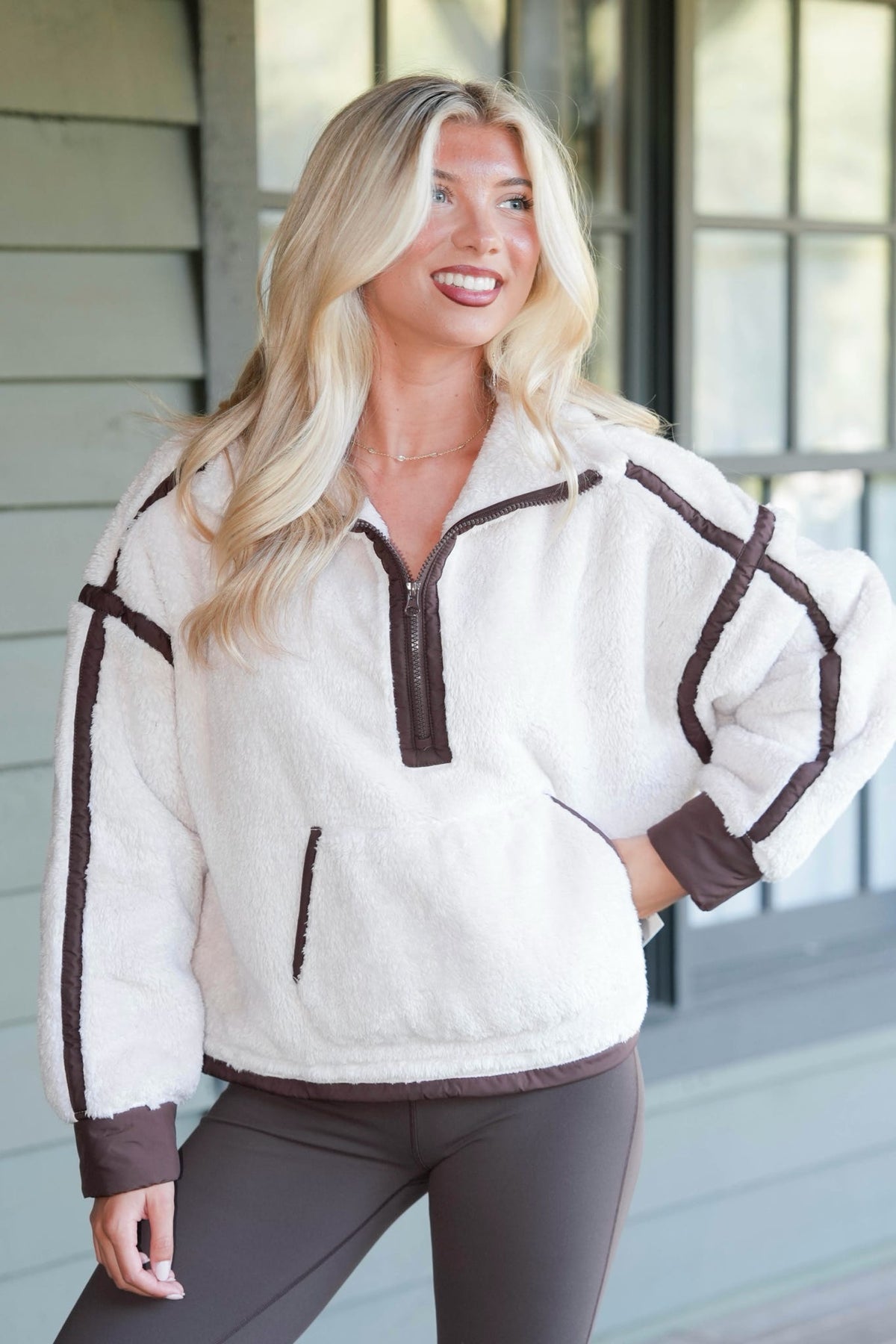 A woman wearing a white sherpa half-zip pullover with dark brown trim and matching dark brown leggings, standing in front of a house exterior.