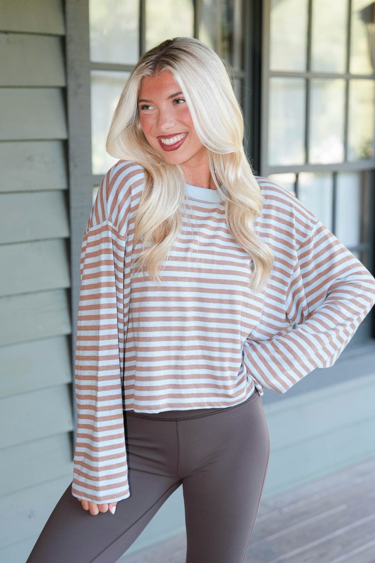 A smiling blonde woman wearing a long-sleeve striped knit top with taupe and white horizontal stripes, paired with dark gray leggings, standing on a porch.