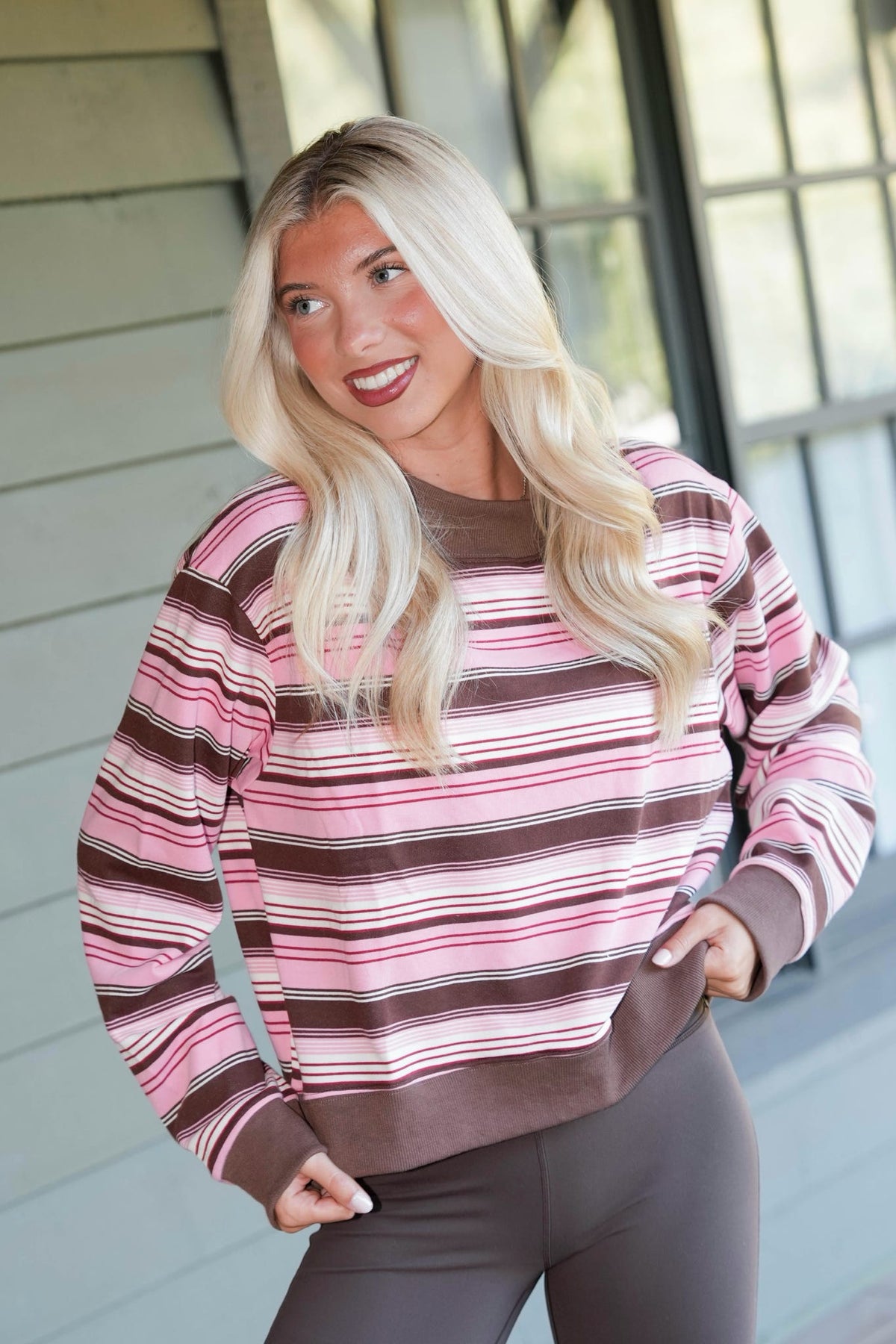 A woman with long blonde hair wearing a pink, brown, and white striped long-sleeve sweatshirt and dark gray leggings, standing in front of a house exterior.