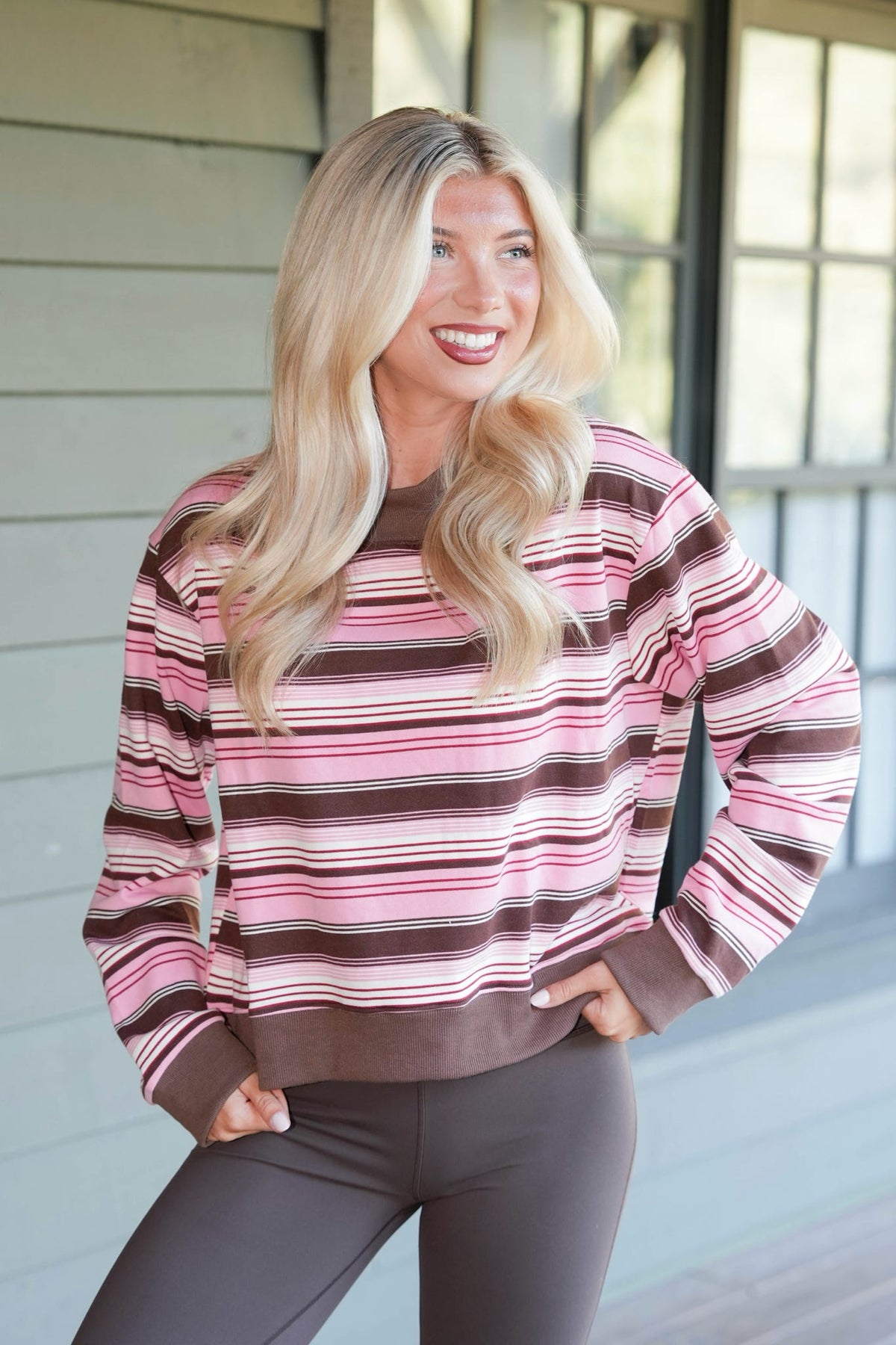 A woman with long blonde hair wearing a pink, brown, and white striped long-sleeve sweatshirt and dark gray leggings, posing on a porch with a light gray wood-paneled wall in the background.