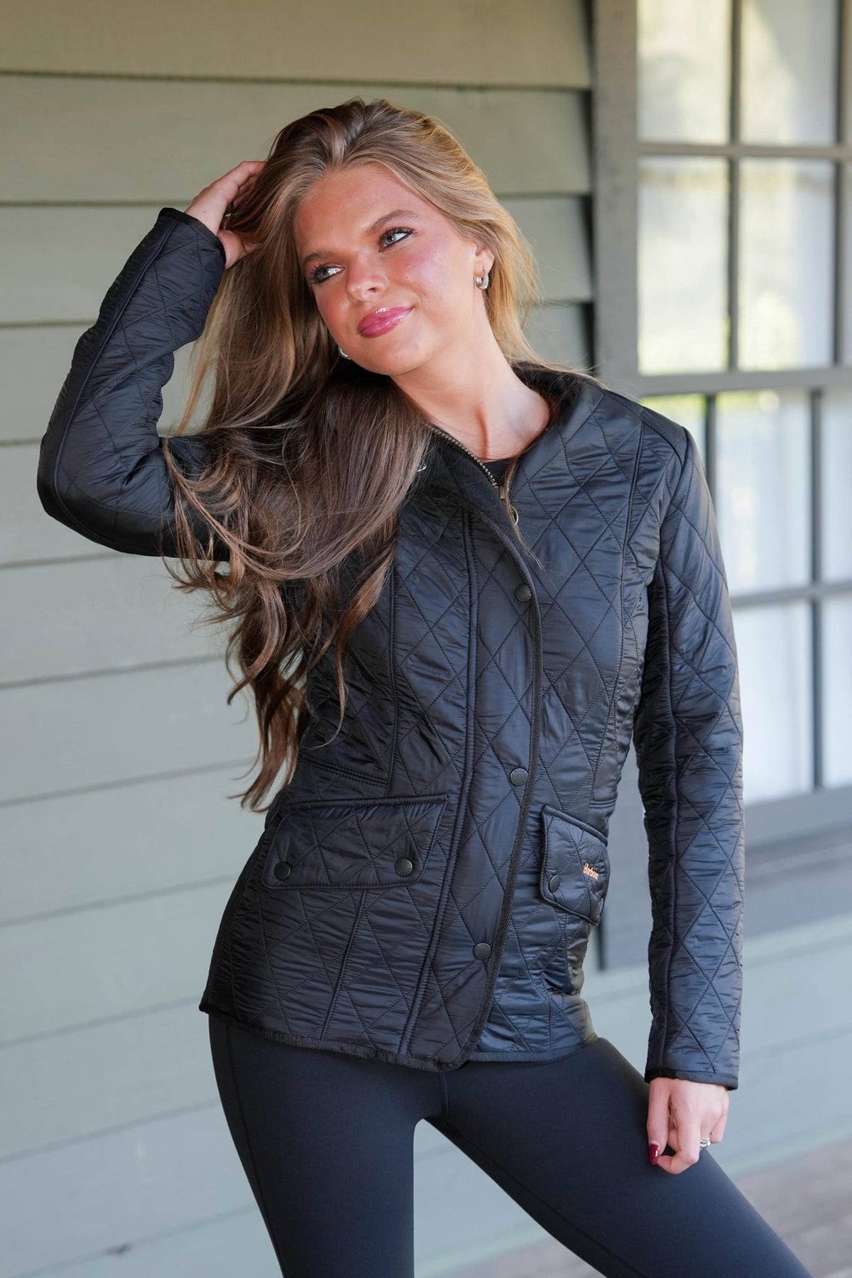 A woman wearing a black quilted jacket and black leggings, posing on a porch with a light gray wood-paneled wall and a window in the background.