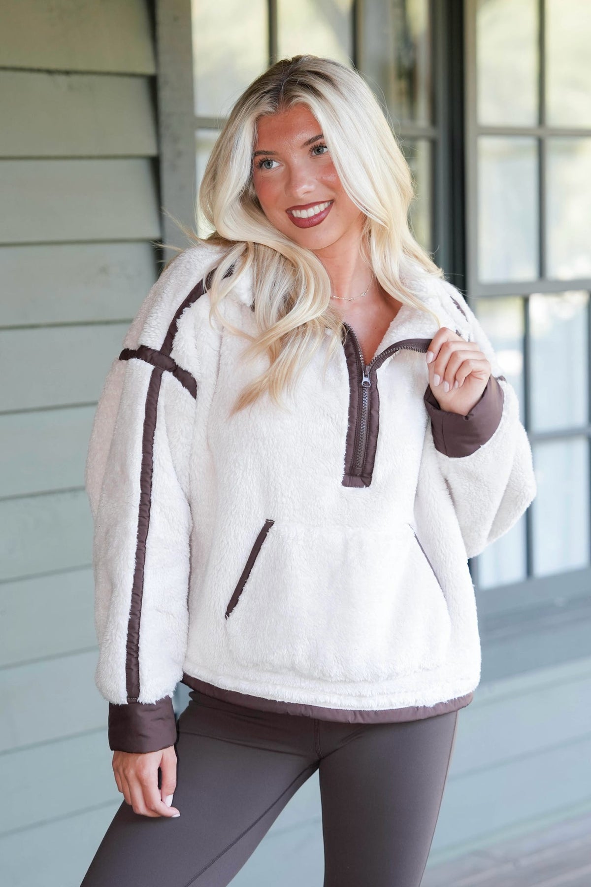 A blonde woman wearing a white sherpa fleece quarter-zip pullover with brown trim, standing in front of a light gray house exterior.