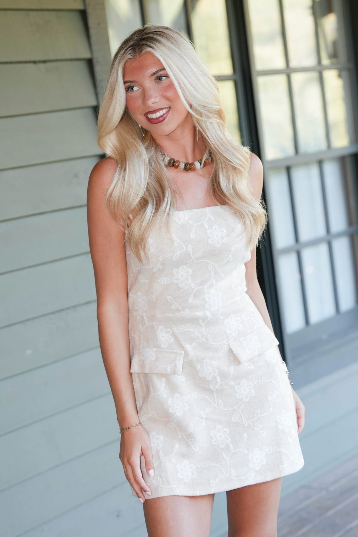 A blonde woman wearing a strapless cream-colored lace mini dress, standing in front of a light gray siding wall and a window.