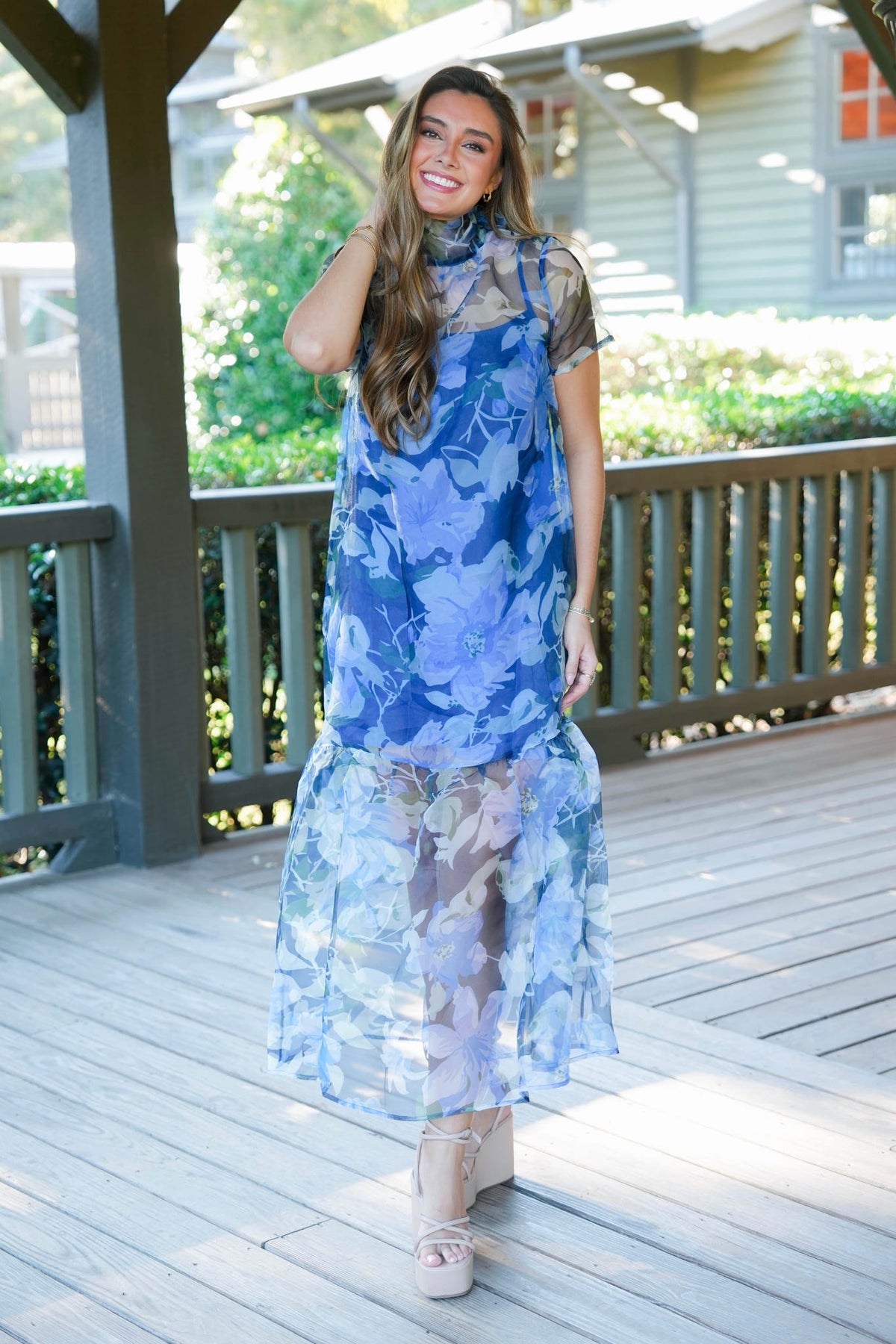 A woman modeling a sheer blue floral midi dress with a high neck and short sleeves, standing on a wooden deck porch.