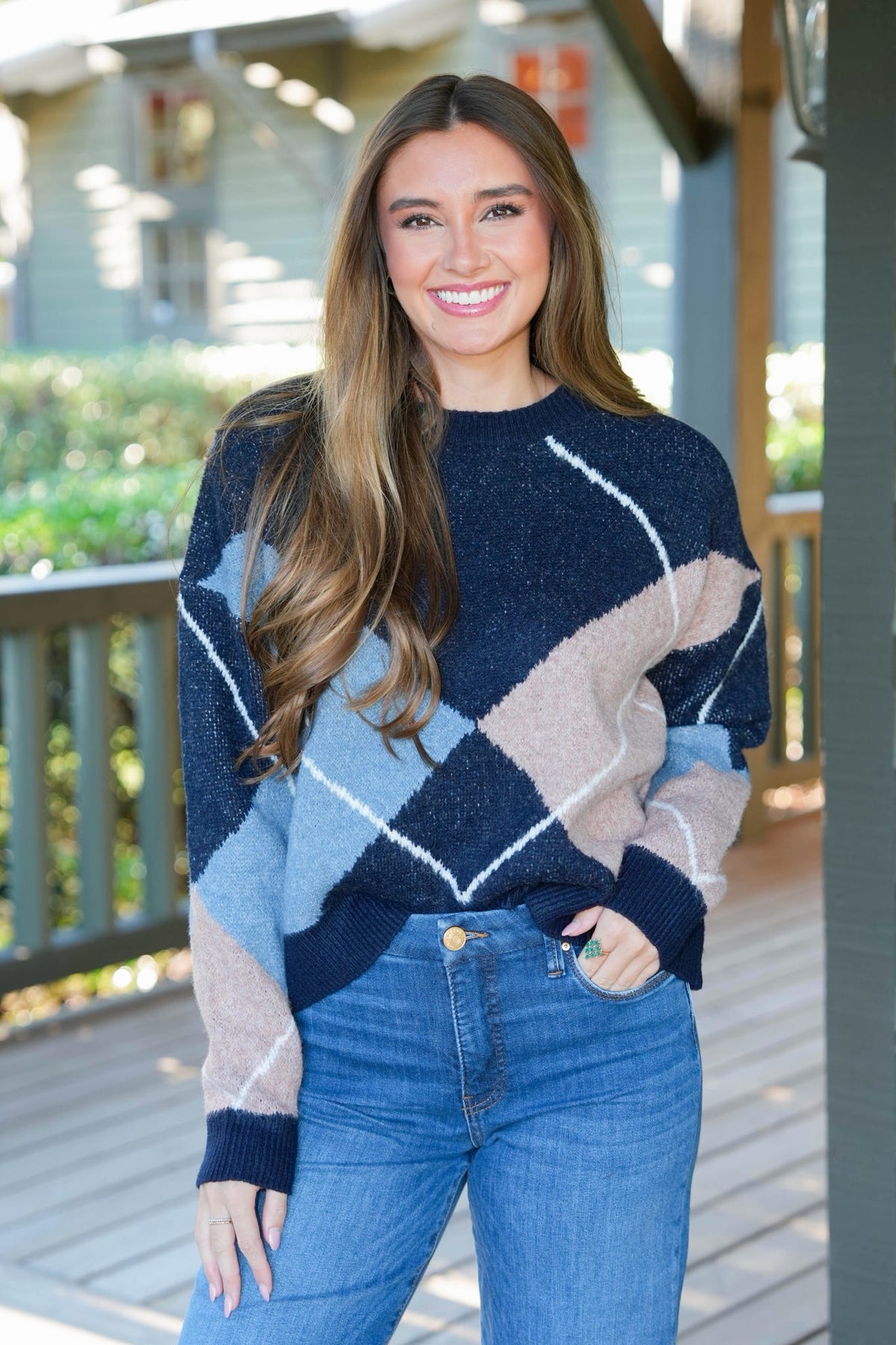 A woman wearing a fuzzy argyle sweater in navy, light blue, and beige with white lines, paired with blue jeans, standing on a wooden porch.