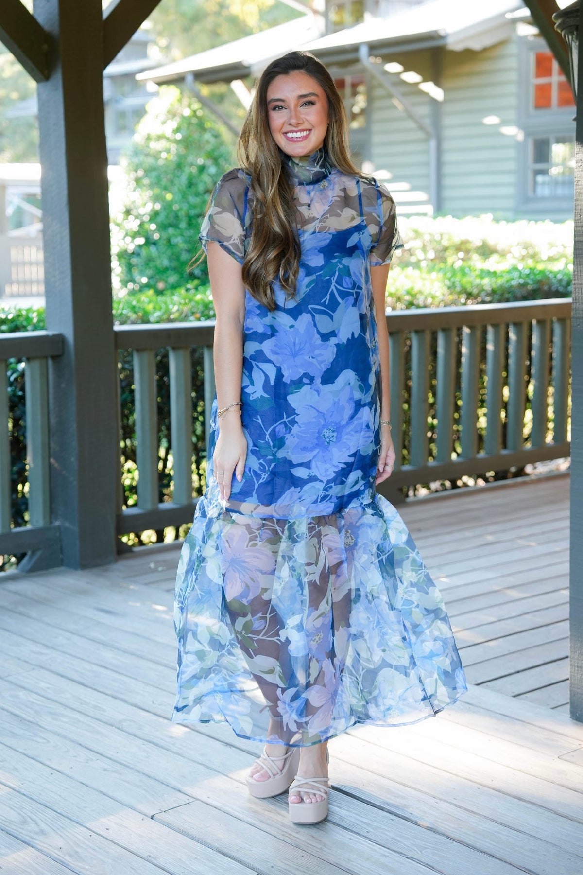 A woman modeling a blue floral midi dress with sheer mesh sleeves and a sheer layered skirt, standing on a wooden deck under a porch.
