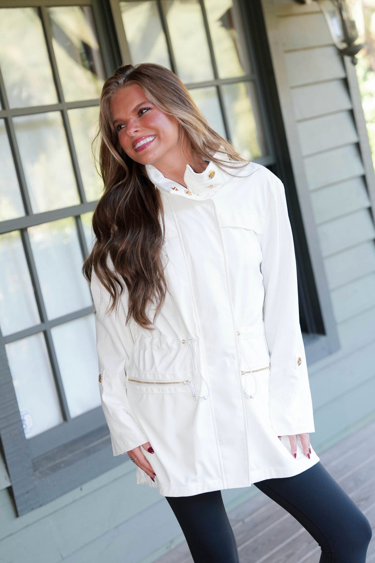 A woman wearing a cream-colored hooded jacket with gold-tone snap buttons and flap pockets, posing on a porch in front of a gray house.