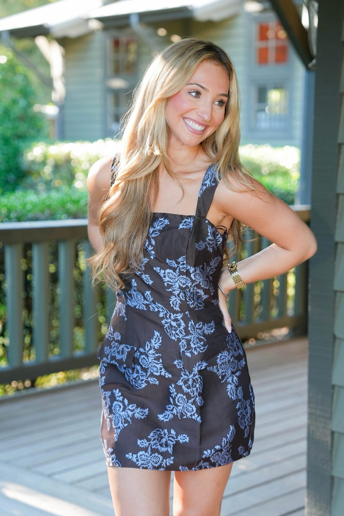 A woman posing on a wooden deck wearing a black mini dress with a light blue floral pattern.