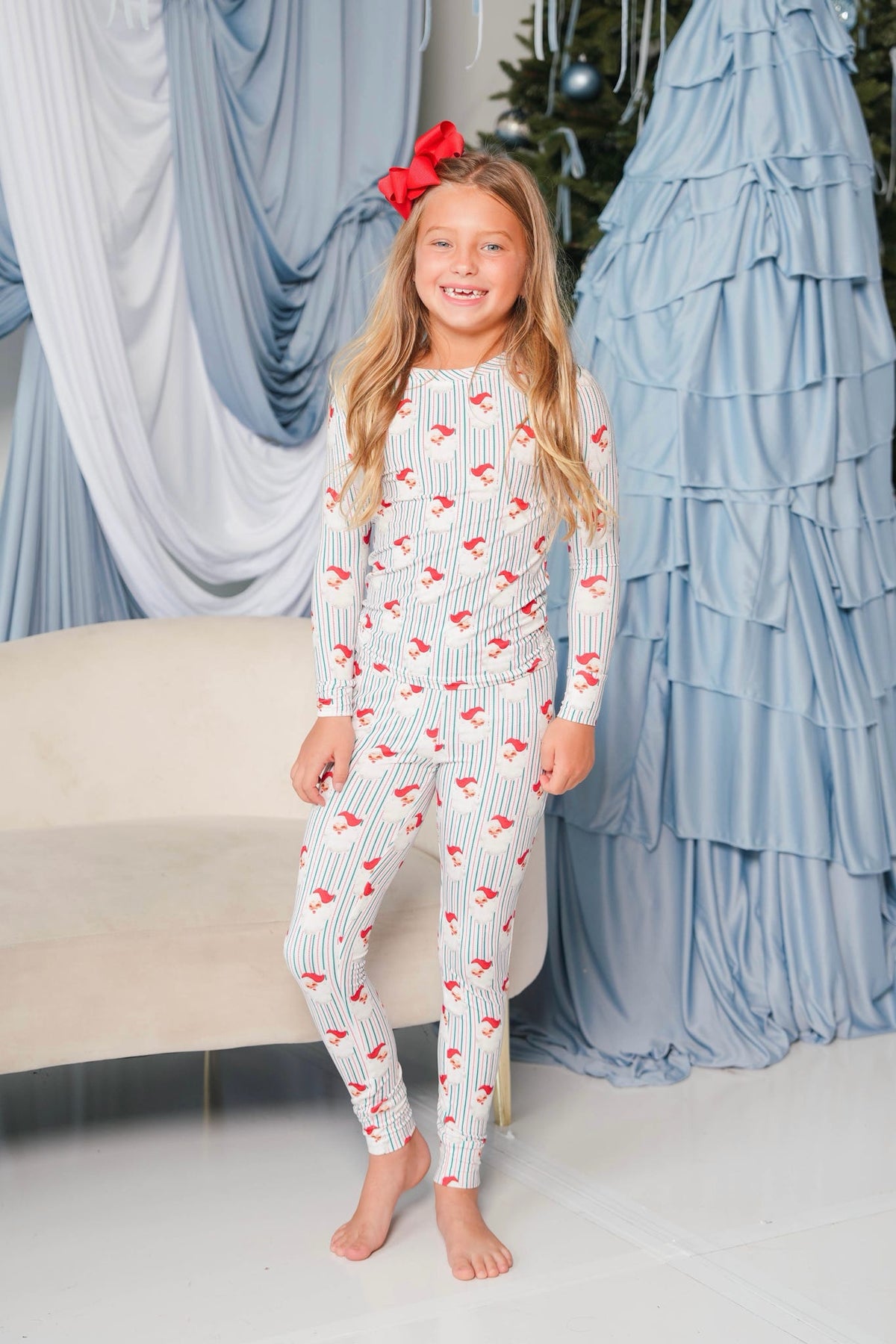 A young girl stands barefoot indoors wearing a long-sleeve, full-length white pajama set with thin vertical gray stripes and a repeating red bird print. She has a red bow in her hair.