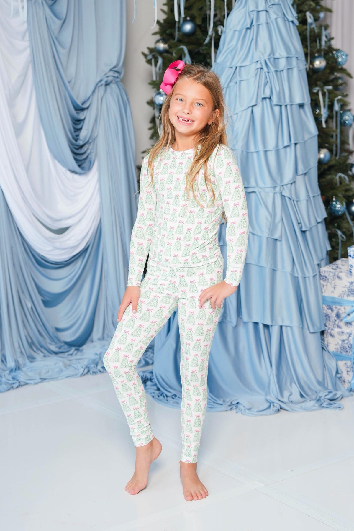 A young girl stands barefoot in a festive holiday setting, wearing a long-sleeve and matching pants pajama set with a light mint green and white geometric pattern. The background features draped light blue fabric, a decorated Christmas tree, and blue ruffled drapery.