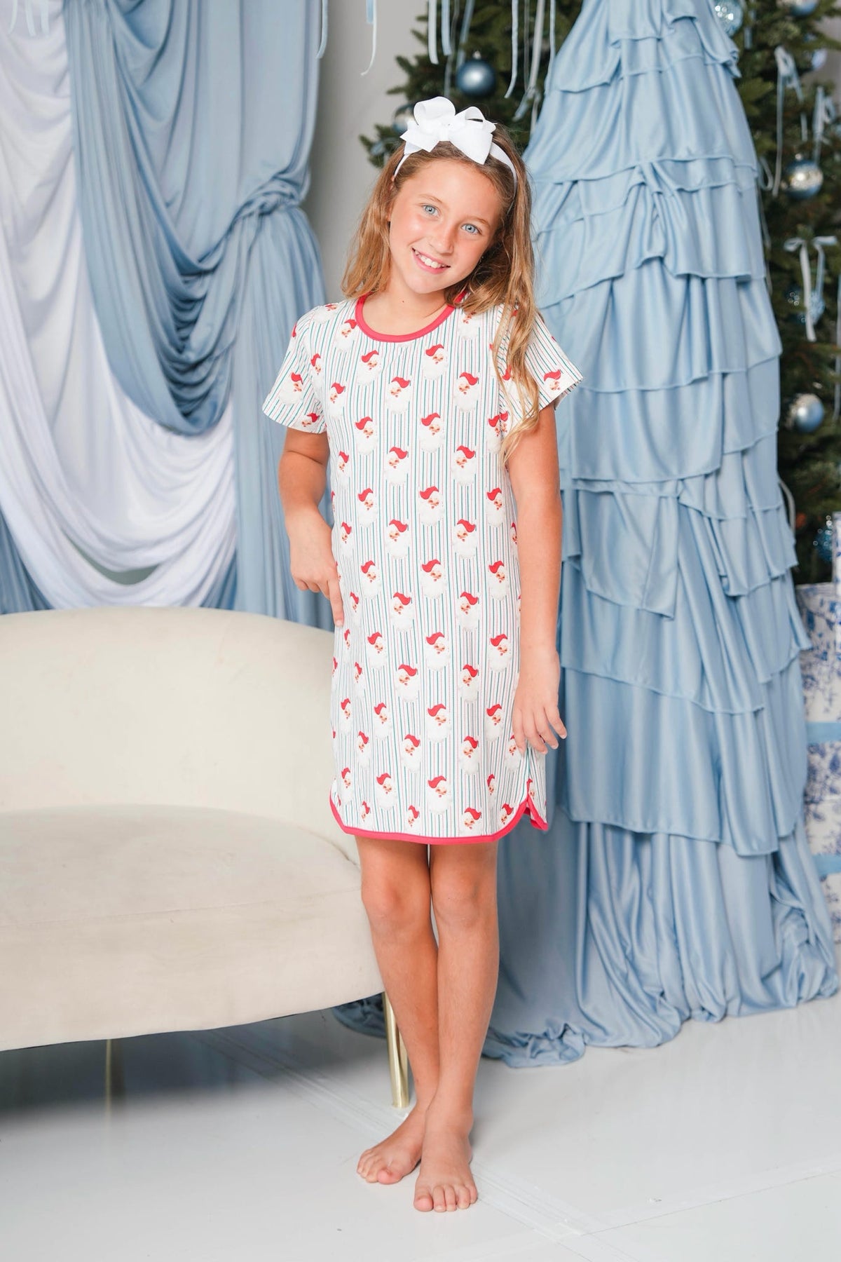 A young girl stands barefoot in a holiday-themed room, wearing a short-sleeve white nightgown with red pinstripes and a red bow pattern, along with a white bow headband.