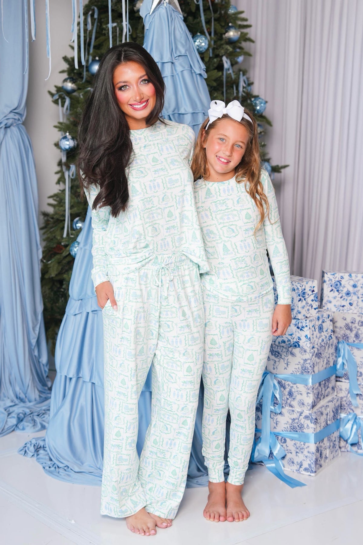 A woman and a young girl wearing matching light blue and white patterned long-sleeve pajamas, standing barefoot in front of a decorated Christmas tree with blue drapes and wrapped presents.
