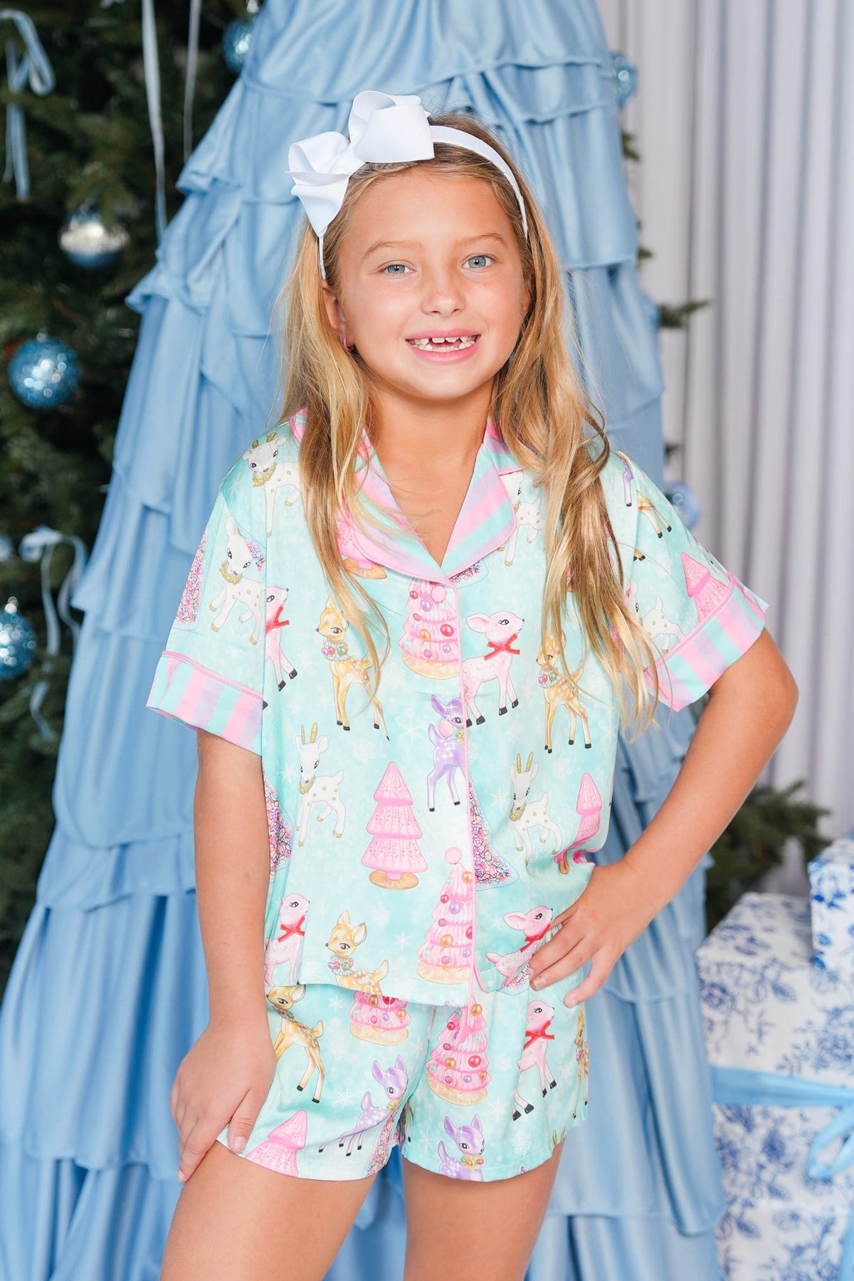 A young girl with blonde hair wearing a light blue and pink unicorn-themed pajama set, featuring a short-sleeve button-up top and matching shorts. She is posing in front of a decorated Christmas tree and a blue ruffled backdrop.