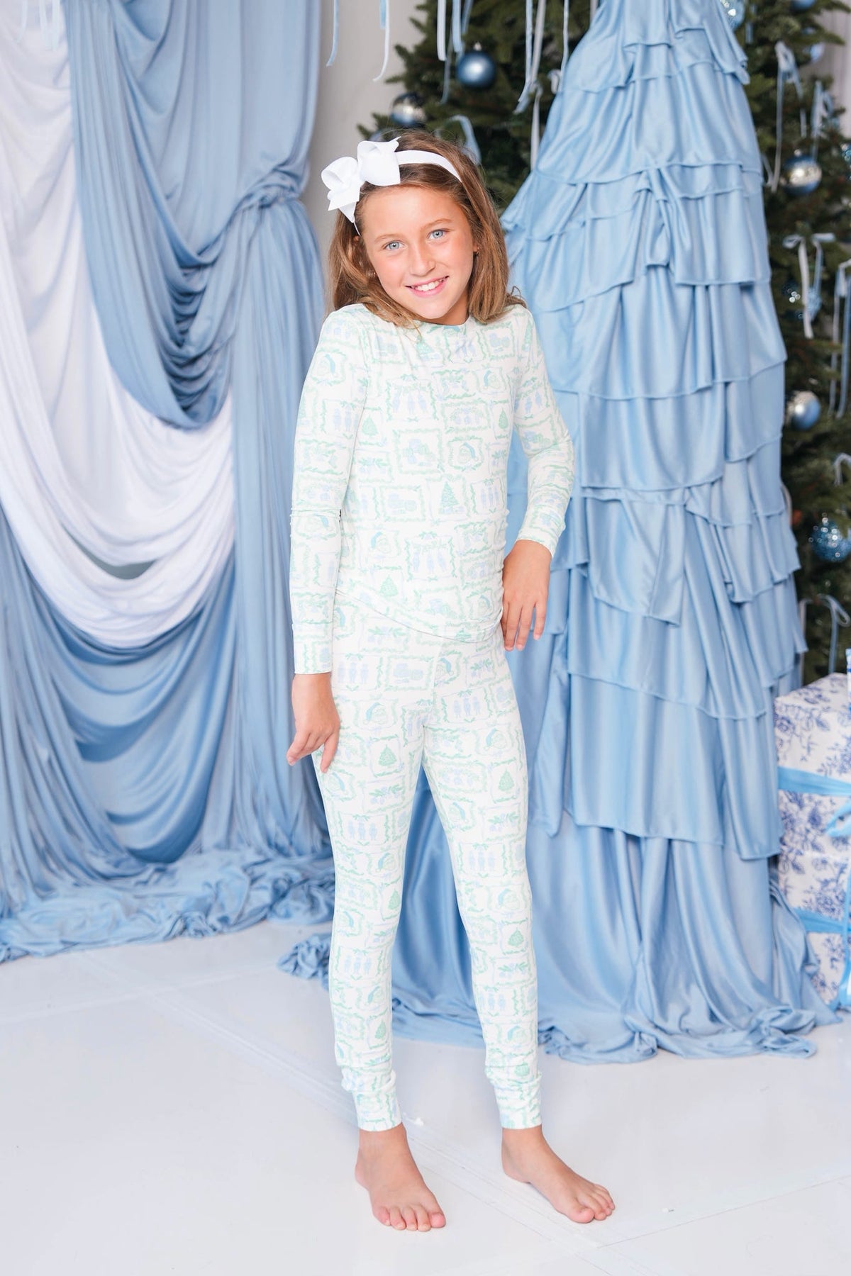 A young girl stands barefoot in a festive studio setting, wearing a light blue and white patterned long-sleeve pajama top and matching pants, with a white bow headband. The background features draped light blue fabric, a decorated Christmas tree, and a ruffled blue backdrop.
