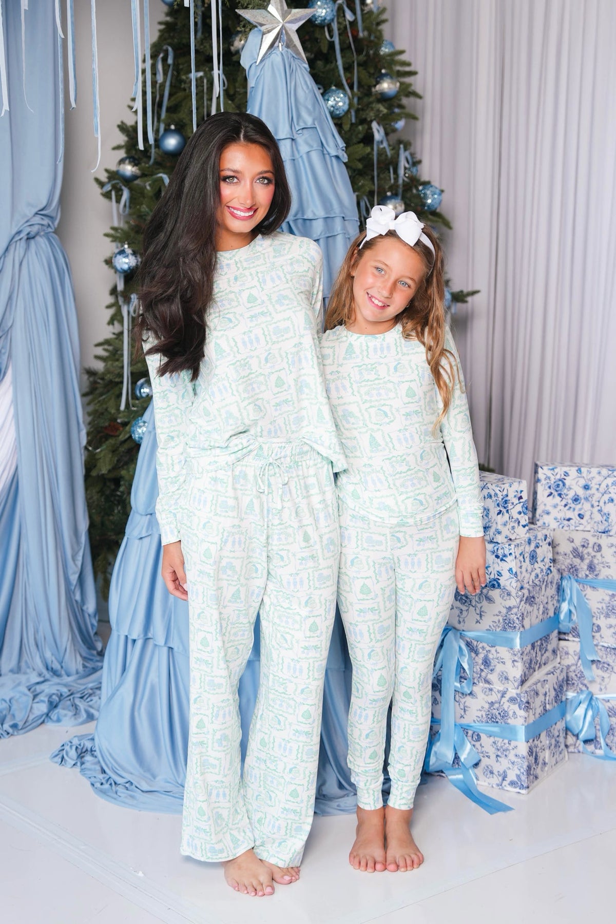 A woman and a young girl wearing matching light blue and white patterned pajama sets, standing barefoot in front of a decorated Christmas tree with blue drapery and wrapped presents.