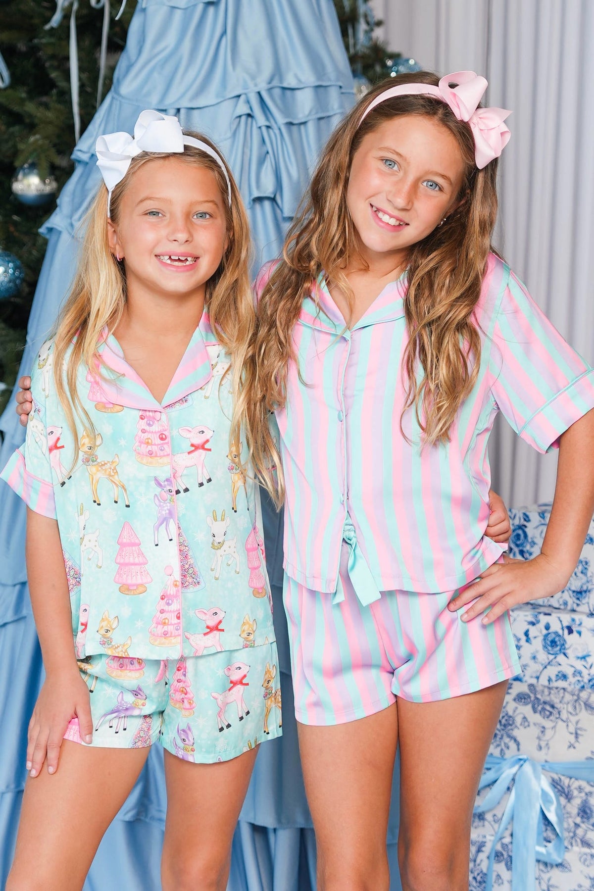 Two young girls wearing matching pastel pajama sets, standing in front of a decorated Christmas tree. The girl on the left wears a light blue pajama set with pink and white snowman prints, and the girl on the right wears a pink and light blue striped pajama set with a matching headband.