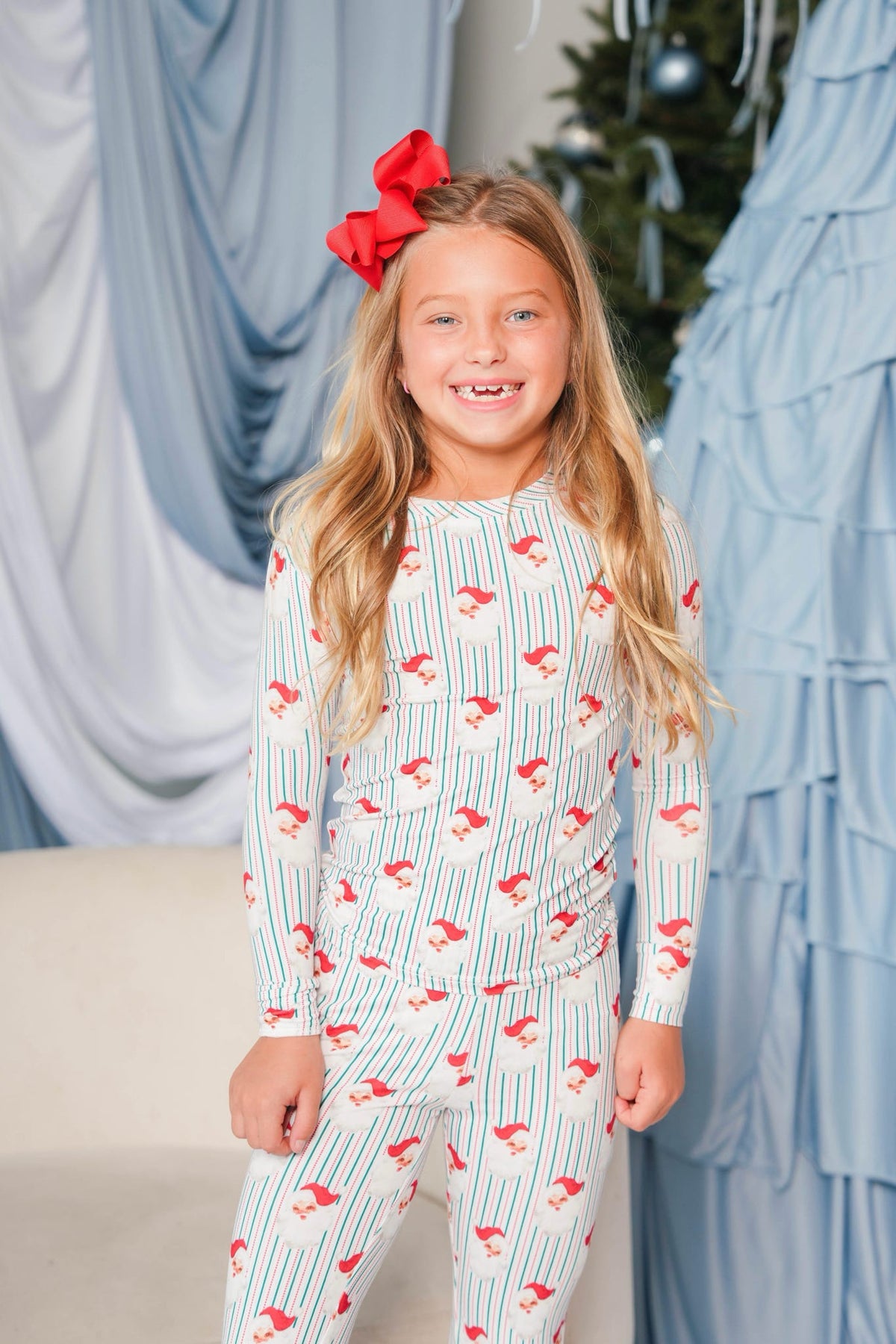A young girl with long blonde hair wearing a red bow headband and a white long-sleeve pajama set with thin blue vertical stripes and small red bird prints, standing in front of a decorated holiday backdrop with blue drapes and a Christmas tree.