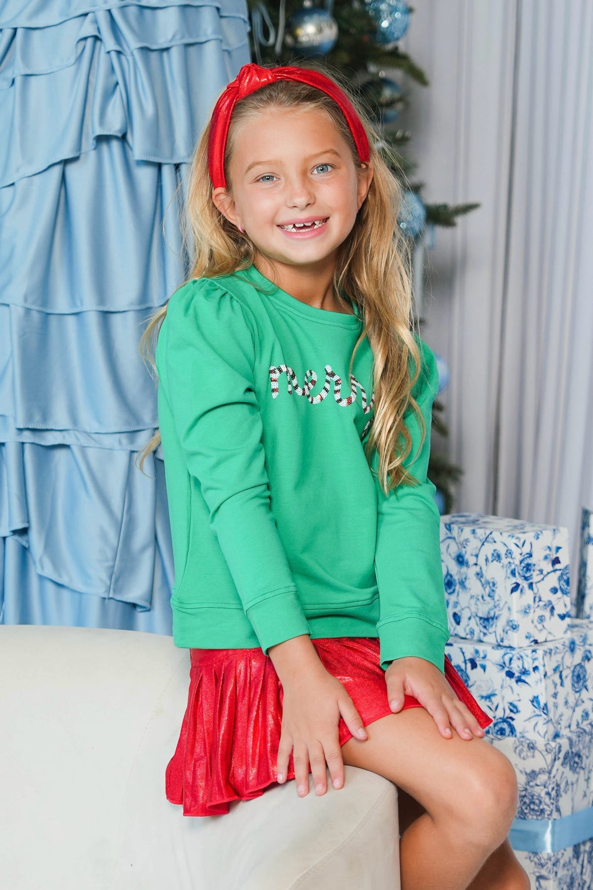 A young girl with blonde hair wearing a bright green long-sleeve top with a rhinestone chest graphic and a shiny red pleated skirt, sitting on a white sofa in a decorated holiday setting.