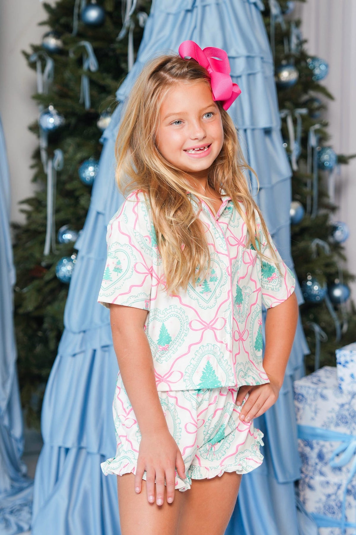 A young girl with a large pink bow in her hair stands in front of a decorated Christmas tree wearing a matching two-piece pajama set with a pink, green, and white pattern. The set includes a short-sleeve button-up top and matching shorts.