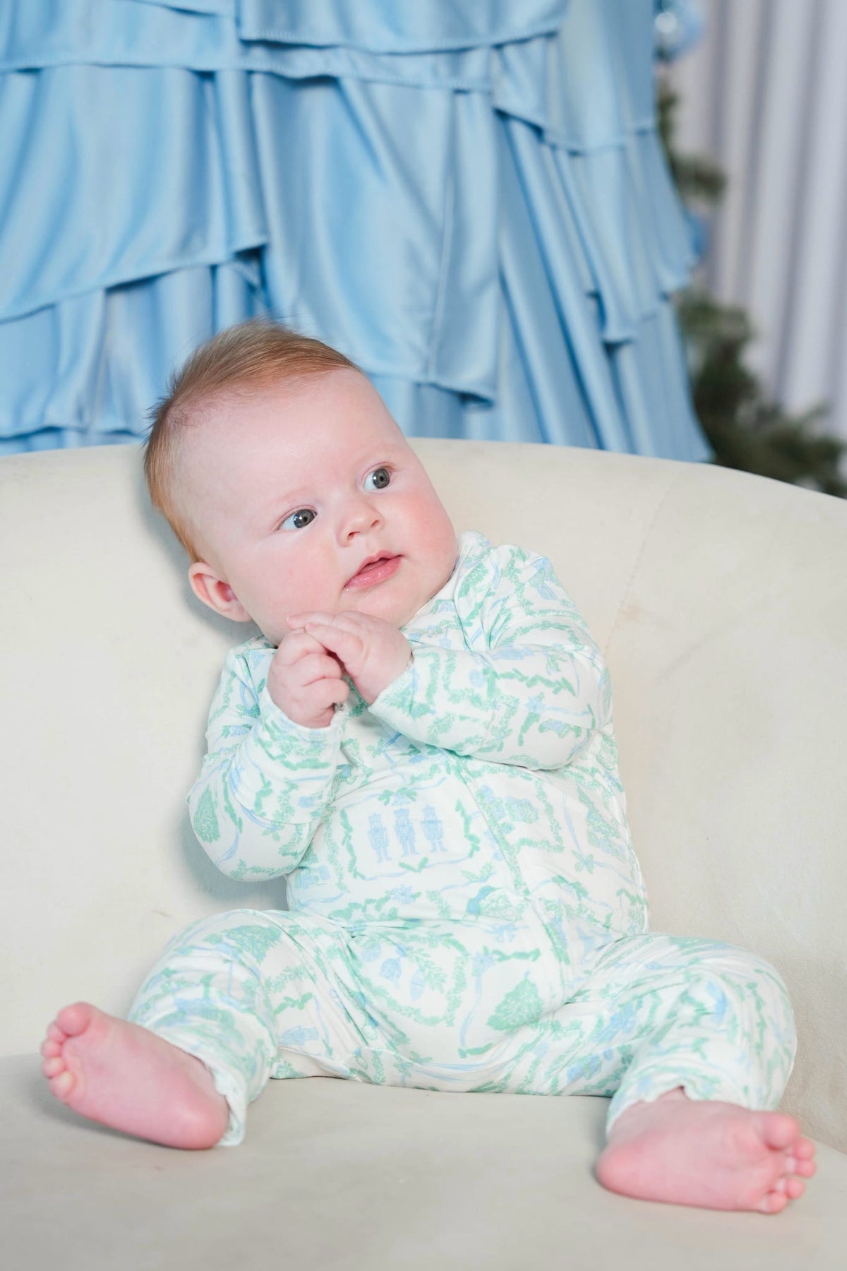A baby sitting on a cream-colored chair, wearing a light green and white patterned long-sleeve footed onesie. The background features a light blue ruffled curtain.