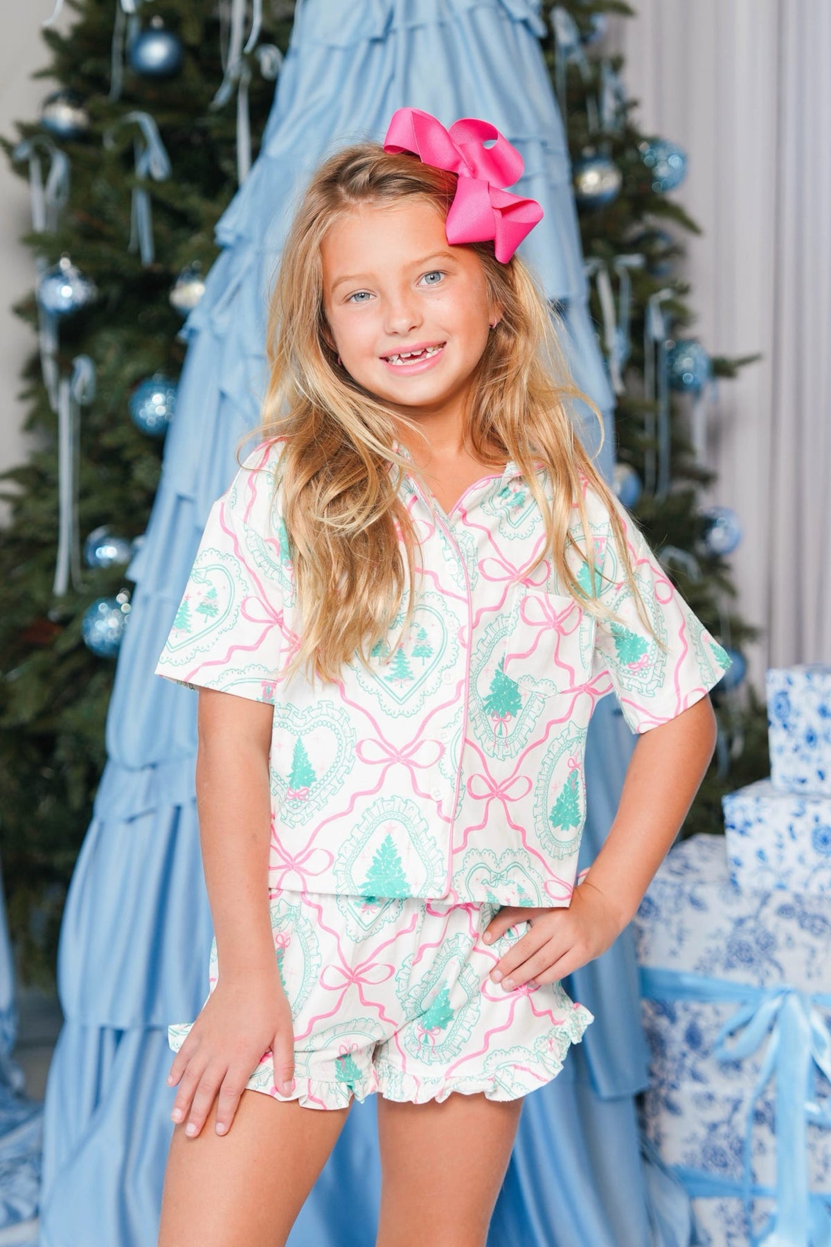A young girl with a large pink bow in her hair stands in front of a decorated Christmas tree wearing a matching short-sleeve button-up shirt and ruffled shorts pajama set. The pajama set features a white background with a pink and mint green geometric pattern.