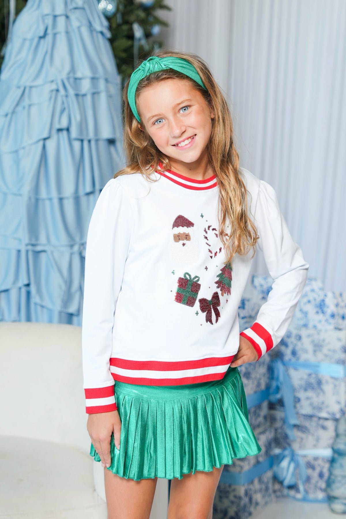 A young girl wearing a white Christmas-themed long-sleeve sweatshirt with red and white striped trim and a festive graphic print, paired with a shiny green pleated skirt and a matching green headband.