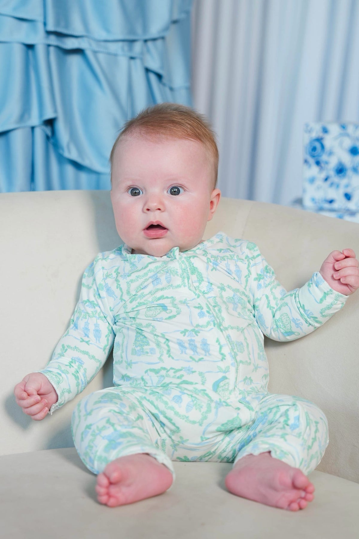A baby sitting on a beige sofa wearing a light green and white patterned long-sleeve footless onesie, with blue curtains in the background.