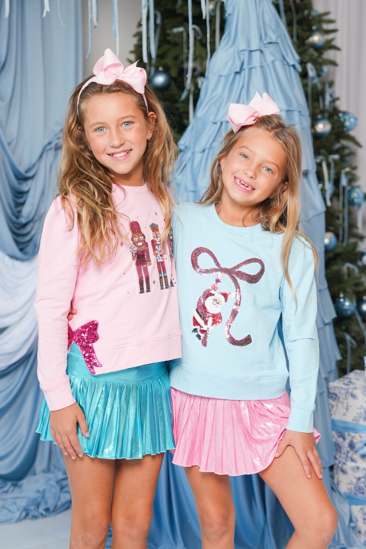Two young girls posing in matching holiday-themed outfits. The girl on the left wears a pink long-sleeve shirt with a nutcracker graphic, a pink bow headband, and a shiny teal pleated skirt.