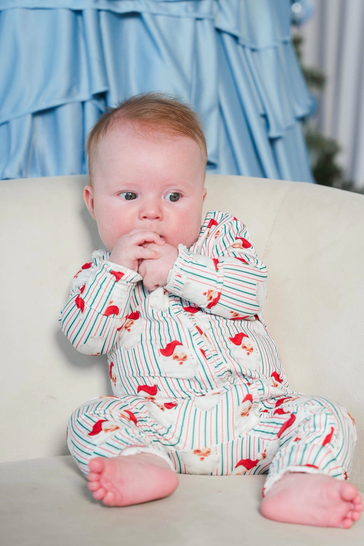 A baby sitting on a cream-colored sofa wearing a white long-sleeve footless onesie with thin green stripes and red bird prints, with a soft blue ruffled backdrop behind.