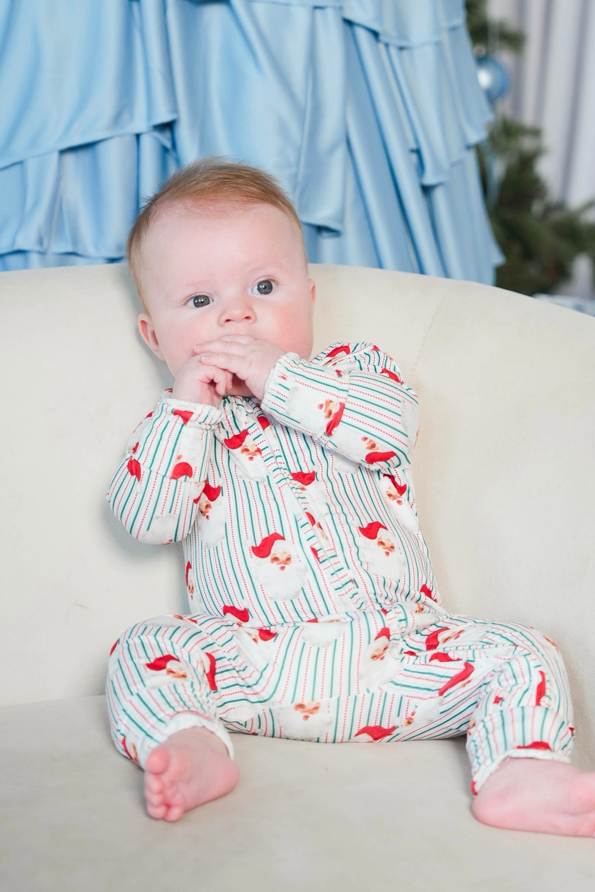 A baby sitting on a cream-colored couch wearing a long-sleeve footless pajama romper with a white base, green vertical stripes, and red bird prints.