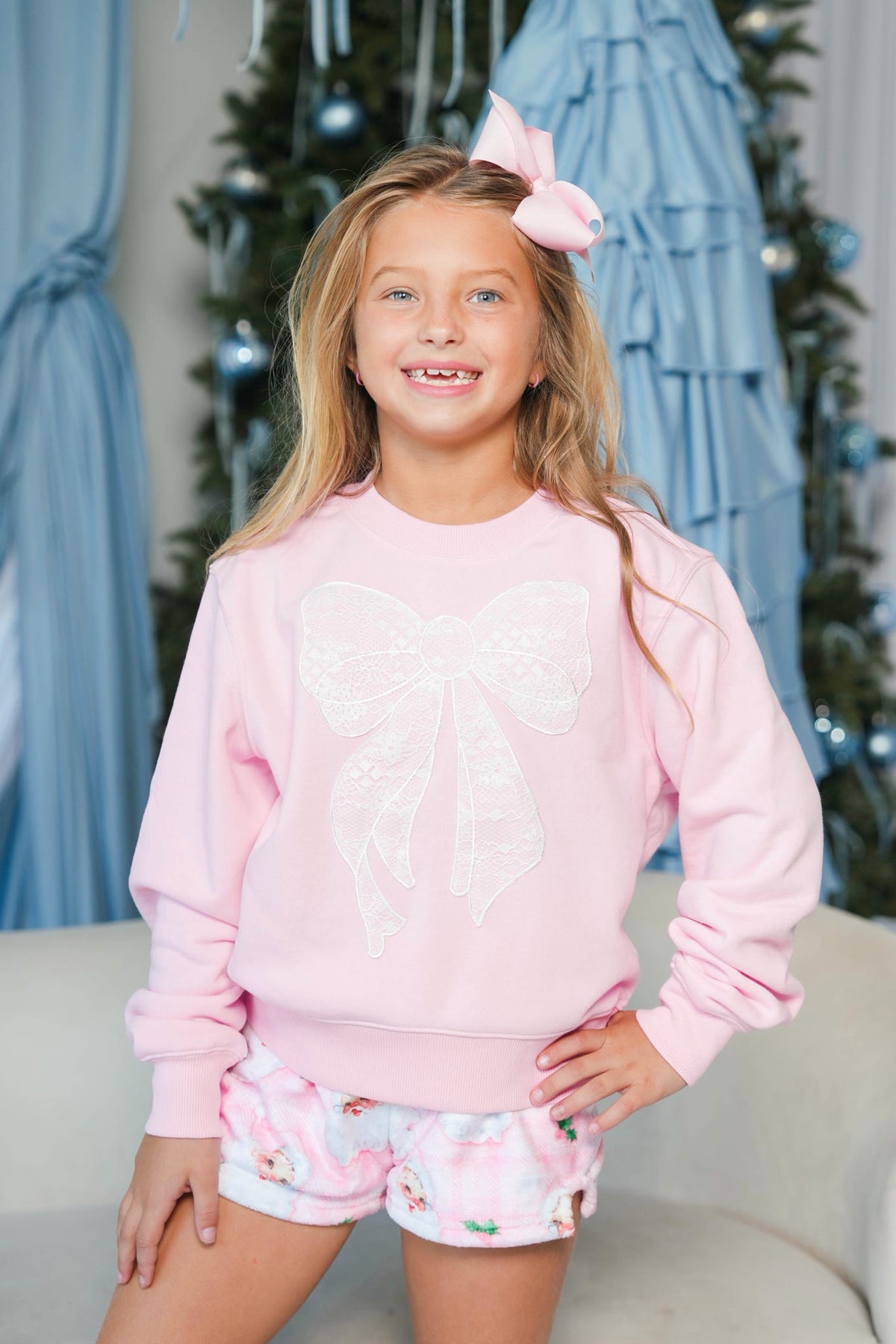 A young girl with blonde hair wearing a light pink crewneck sweatshirt with a large white bow applique on the front, paired with matching pink shorts, posing in front of a decorated Christmas tree.