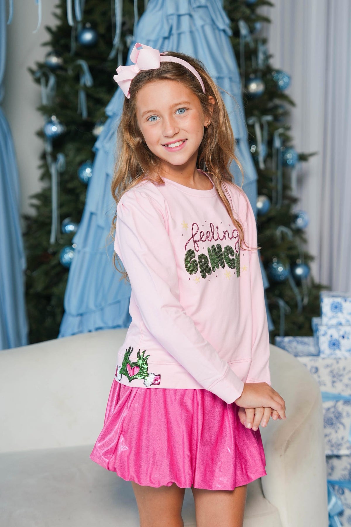 A young girl wearing a pink long-sleeve graphic sweatshirt and a bright pink skirt, standing in a decorated holiday setting with a Christmas tree and wrapped gifts in the background.