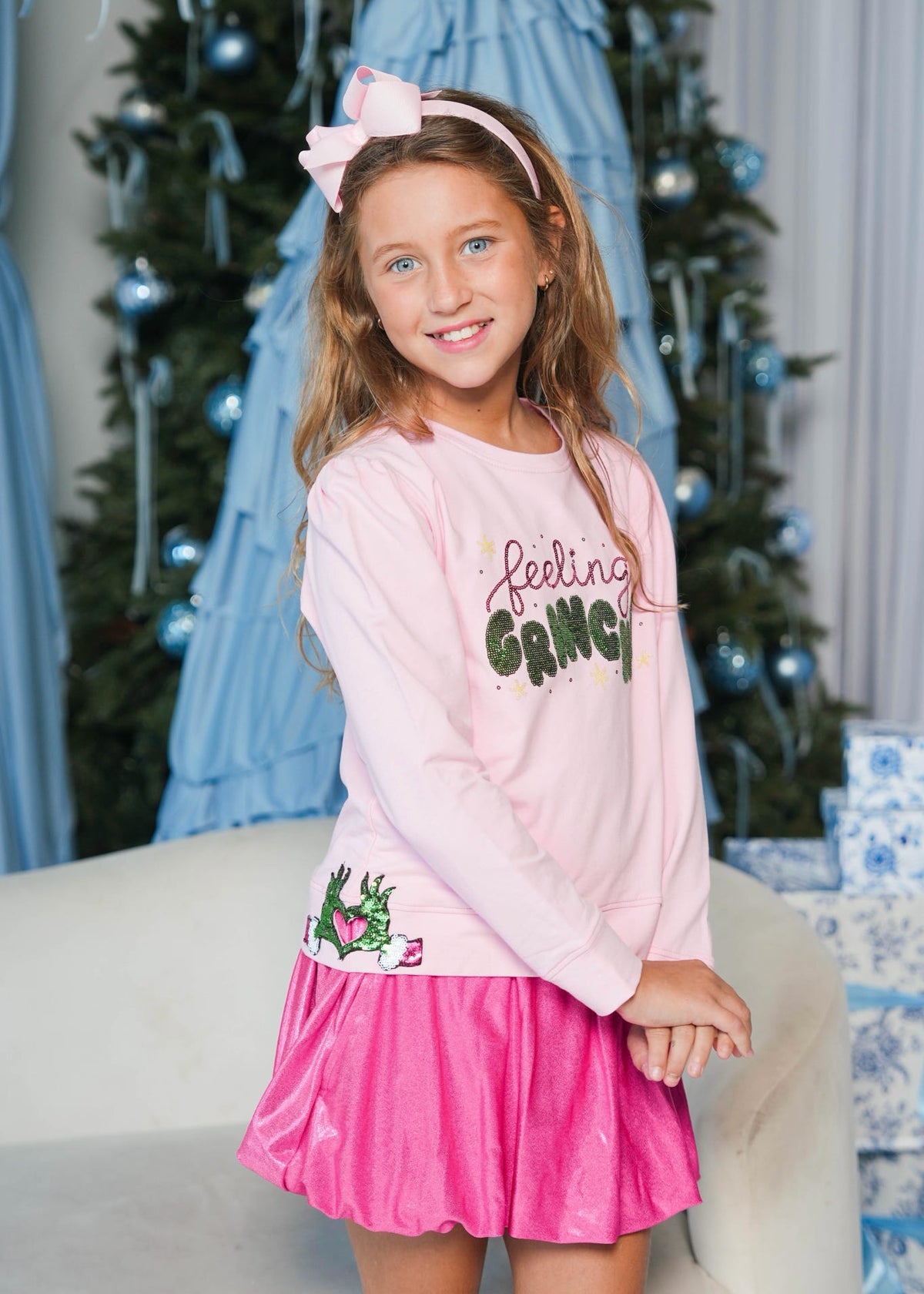 A young girl wearing a pink long-sleeve graphic sweatshirt and a bright pink skirt, standing in a decorated holiday setting with a Christmas tree and wrapped gifts in the background.