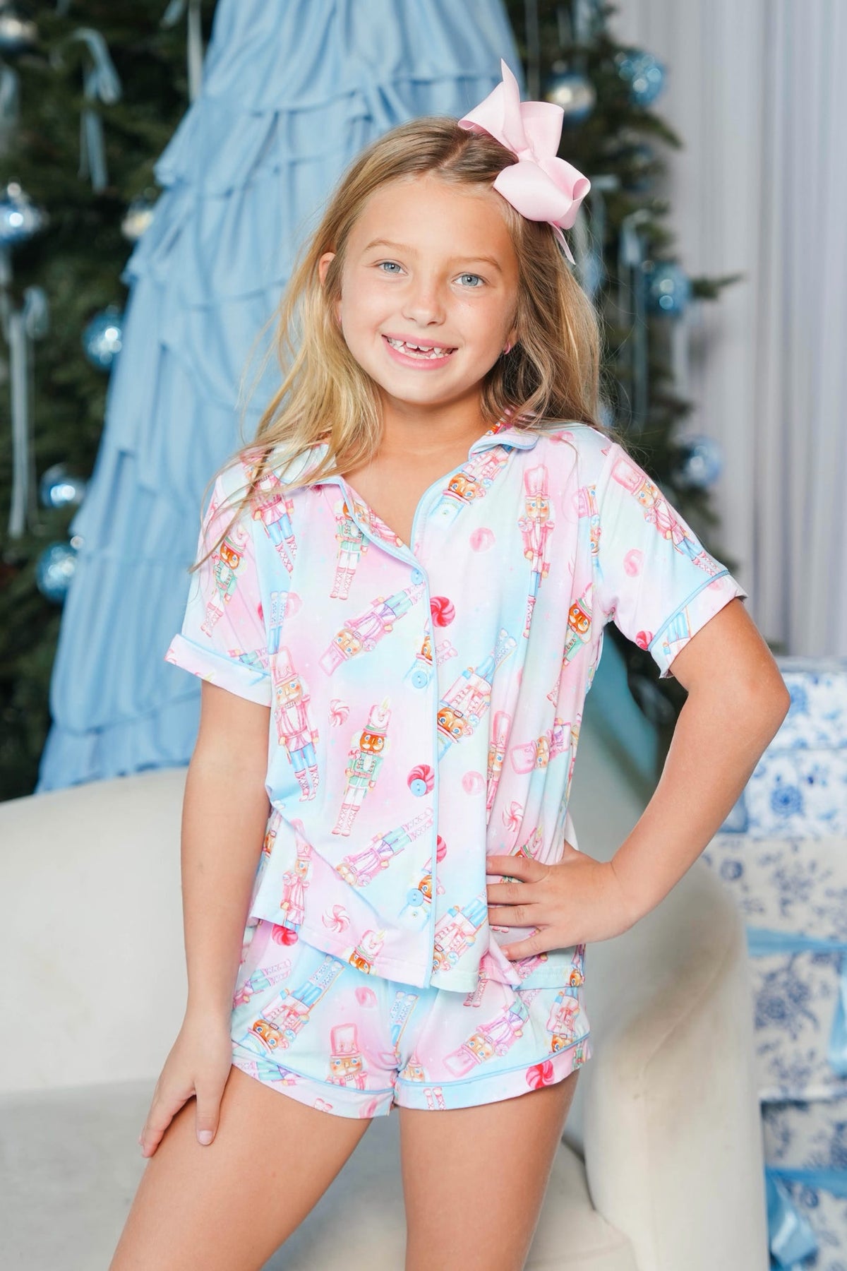 A young girl with blonde hair and a pink bow smiles while wearing a light blue and pink pajama set with a cartoon print, standing in a decorated indoor setting with a Christmas tree in the background.