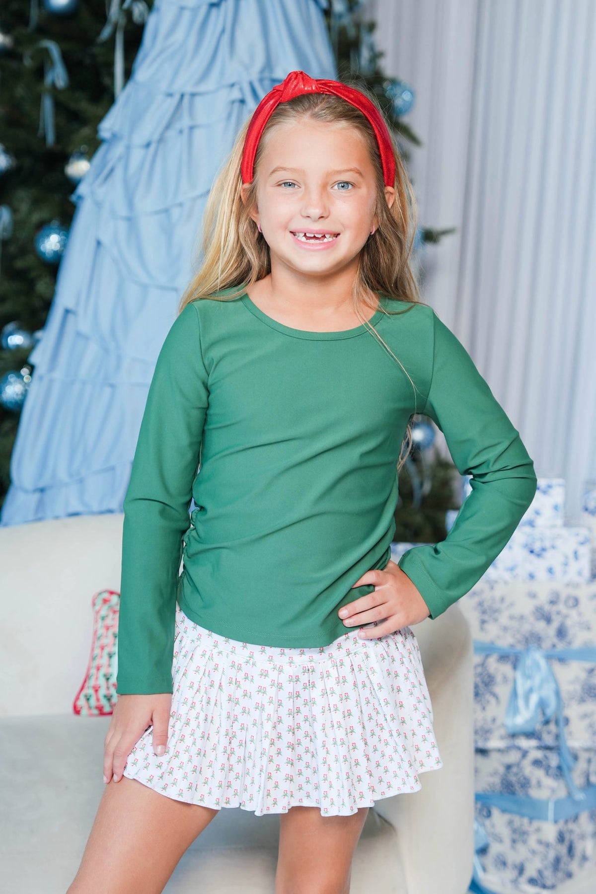 A young girl with blonde hair wearing a green long-sleeve top, a white patterned skirt, and a red headband, posing in front of a decorated Christmas tree and holiday decorations.