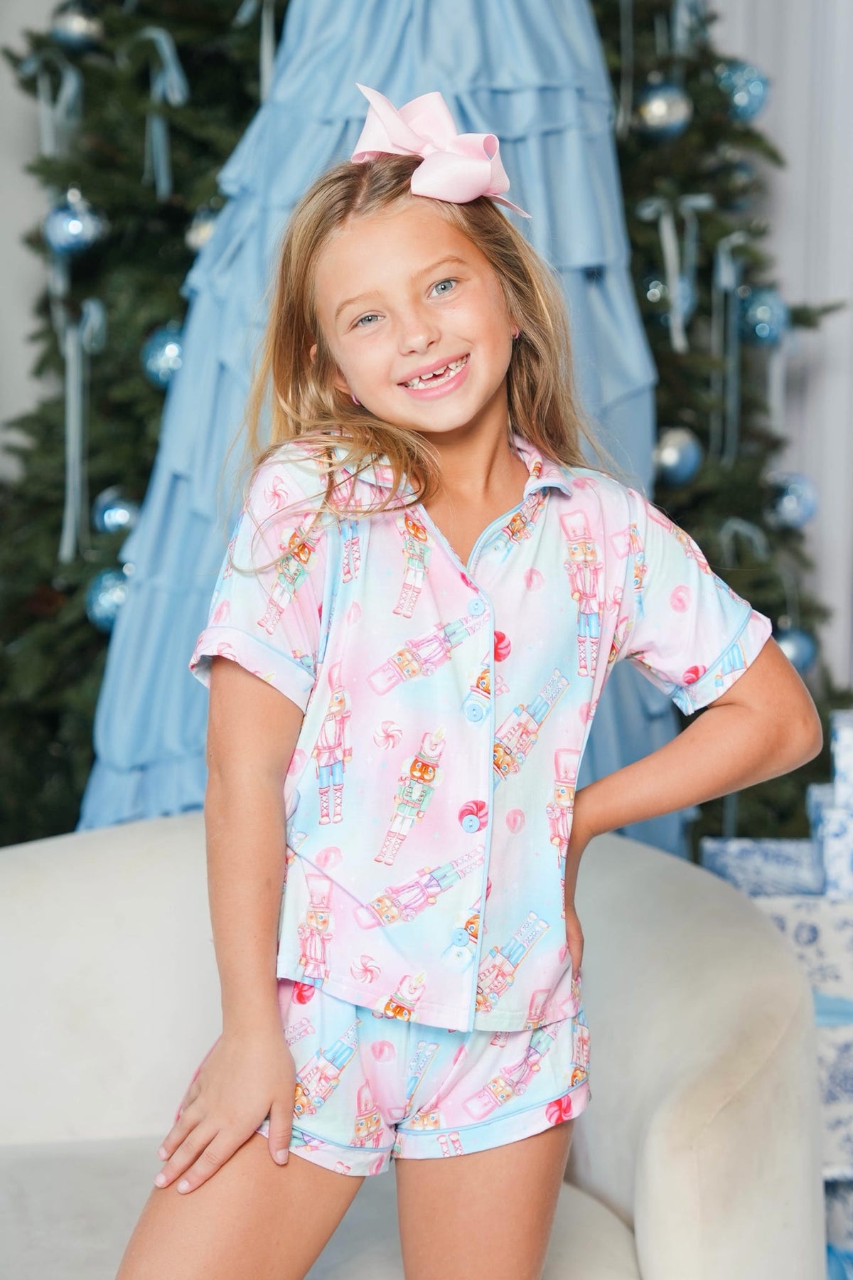 A young girl smiling while wearing a light blue and pink Christmas-themed pajama set with a nutcracker print, posed in front of a decorated Christmas tree and blue fabric backdrop.