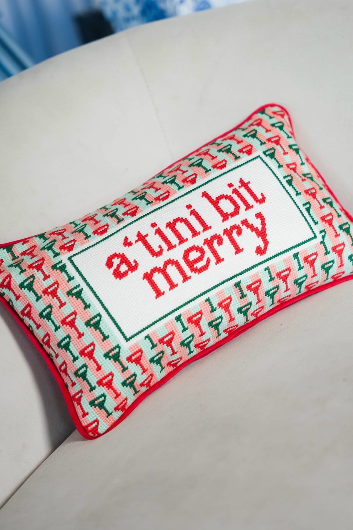 A rectangular decorative pillow with a red and green patterned border and the phrase “a’tini bit merry” embroidered in red inside a white center panel, resting on a light-colored sofa.
