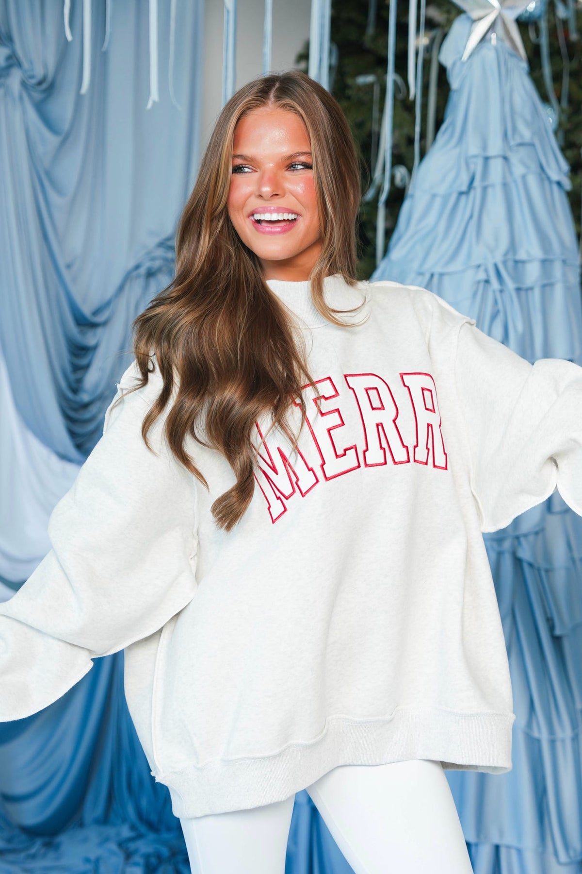 A woman wearing an oversized cream sweatshirt with 'MERR' in red collegiate-style lettering, posed against a blue draped fabric backdrop.
