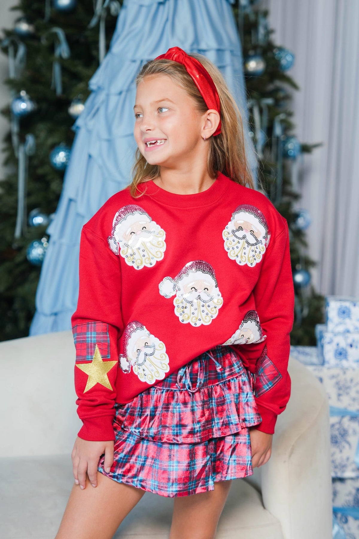 A young girl wearing a festive Christmas sweater and matching plaid skirt, posed in front of a decorated Christmas tree and blue tulle backdrop.
