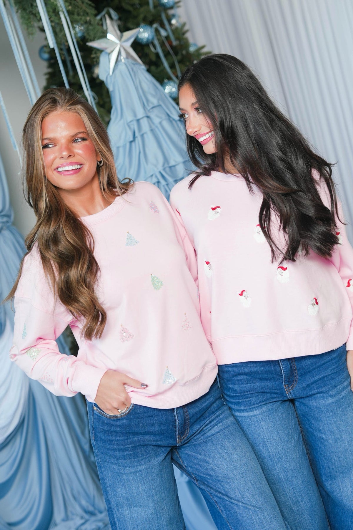 Two women wearing matching light pink crewneck sweaters with small embroidered holiday motifs, paired with blue jeans, standing in front of a decorated Christmas tree and blue draped fabric.